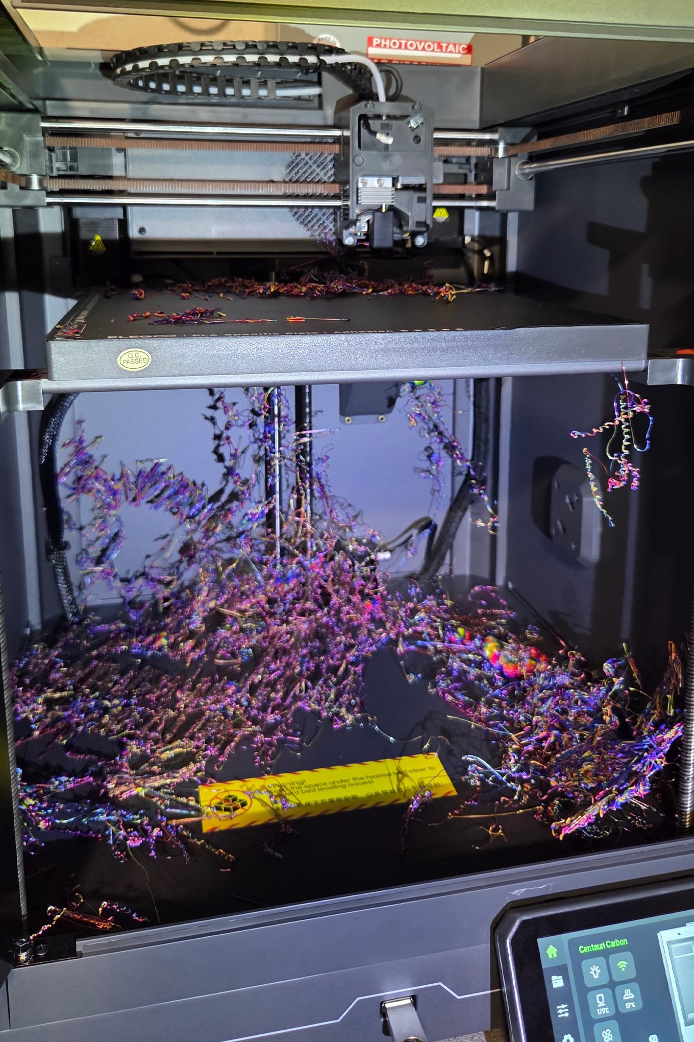 Inside the box of a 3D printer, strings and gobs of rainbow filament are strewn across the bed, hang from the platform inside, and cover its floor.