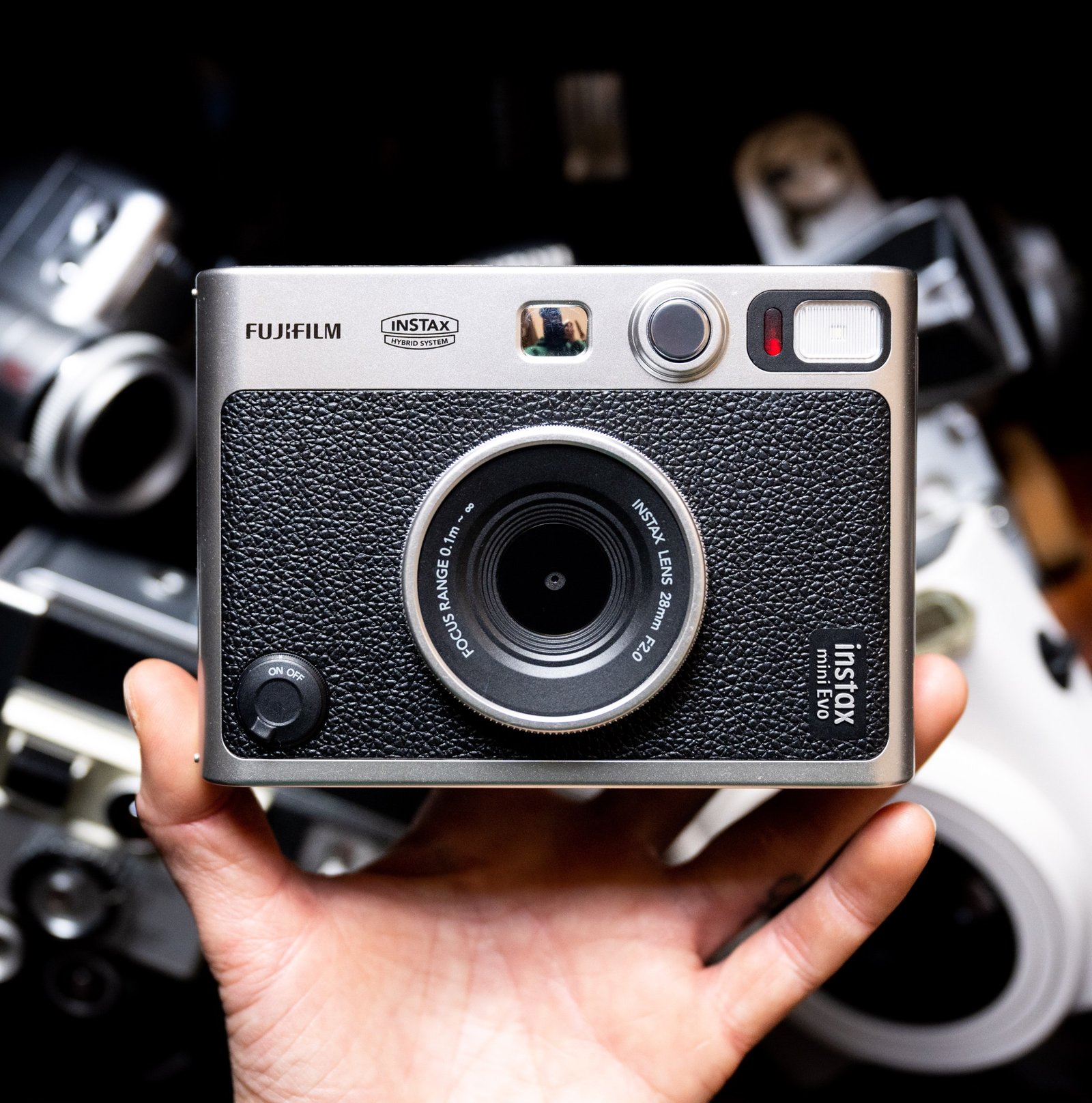 The Instax Mini Evo being held up with a hand against a dark background with cameras that are out of focus.